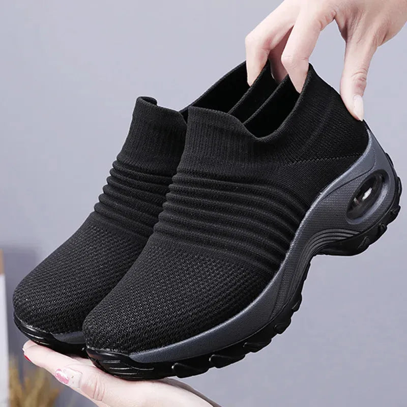 Black slip-on sneakers held by a hand against a gray background