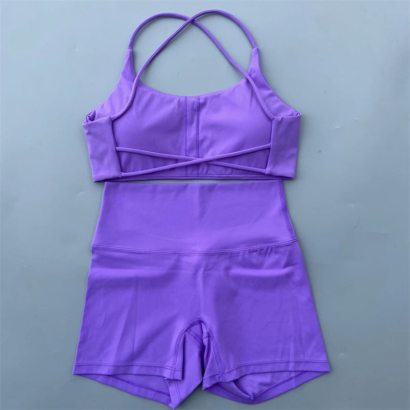 A two-piece purple athletic set consisting of a sports bra top with crossover underbust straps and matching high-waisted shorts, laid flat on a gray background.