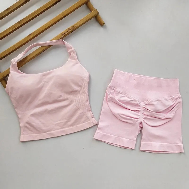 A light pink two-piece activewear set consisting of a halter-style crop top and ruched high-waisted shorts, laid flat on a light gray surface with a wooden slatted prop in the top-left corner.